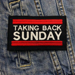May include: A black rectangular patch with the words "TAKING BACK SUNDAY" embroidered in white, framed by two red horizontal stripes. The patch is sewn onto a denim jacket, showcasing a classic style.