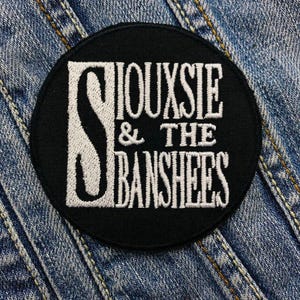 May include: A black, circular patch with white embroidered text. The text reads "SIOUXSIE & THE BANSHEES" in a stylized font. The patch is set against a denim background.