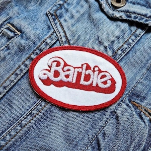 Op de afbeelding: Een geborduurde patch met het woord "Barbie" in witte letters, omrand met rood, op een witte ovale achtergrond. De patch is genaaid op een blauw spijkerjasje.
