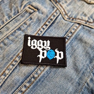 May include: A black rectangular patch featuring the words "Iggy Pop" in white, stylized lettering and a blue sphere. The patch is sewn onto a light blue denim jacket.