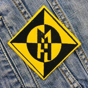 Könnte beinhalten: Ein gelber und schwarzer rautenförmiger Patch mit einem stilisierten "H" in einem Kreis. Der Patch ist aus Stoff und hat einen genähten Rand.