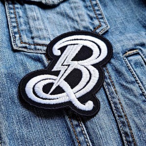 Pode incluir: Patch bordado com uma letra "B" branca estilizada e um design de raio num fundo preto. O patch é costurado numa jaqueta jeans, mostrando seu uso potencial como acessório de vestuário.