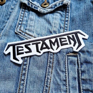 Op de afbeelding: Witte patch met zwarte tekst "TESTAMENT" op een blauw denim jack.