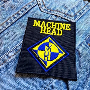 Puede incluir: Parche bordado negro con logotipo Machine Head amarillo y azul. El logotipo presenta una brújula estilizada con una calavera en su interior.