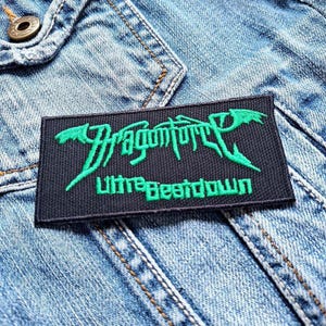 Pode incluir: Um remendo bordado preto e verde com o texto "DragonForce Ultra Beatdown" em um fundo de jeans.