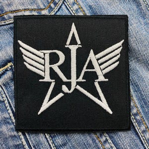 Puede incluir: Parche cuadrado negro con diseño de estrella y alas bordadas en blanco. Las letras "RJA" están centradas dentro de la estrella. El parche está sobre un fondo de mezclilla.