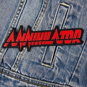 Puede incluir: Un parche bordado rojo y negro con la palabra "Annihilator" en una fuente audaz y estilizada. El parche está cosido en una chaqueta vaquera azul.