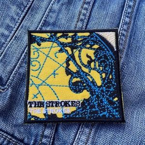 May include: Yellow and blue embroidered patch with the text "The Strokes" and a graphic design of a map.
