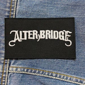 Op de afbeelding: Een zwart rechthoekig embleem met de witte tekst "ALTER BRIDGE" in een gestileerd lettertype. Het embleem is geplaatst op een blauwe denim achtergrond, met zichtbare stiksels.
