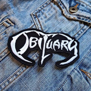 Op de afbeelding: Zwart-wit geborduurde patch met het woord "Obituary" in een gestileerd lettertype. De patch is genaaid op een blauw denim jack.