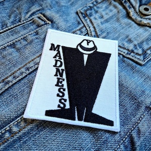 May include: A white rectangular patch with the word "MADNESS" in black capital letters on the left side. A black silhouette of a man wearing a hat is in the center. The patch is on a denim jacket.