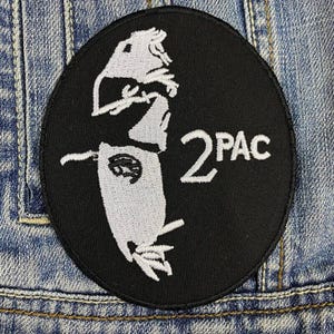Puede incluir: Parche negro, de forma ovalada, con la silueta blanca de un rostro y el texto "2PAC". El parche está cosido a una chaqueta vaquera. Un diseño clásico, adecuado para ropa o accesorios.
