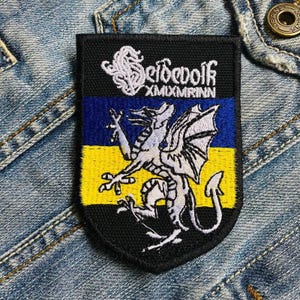 May include: Black embroidered patch with a yellow and blue striped background. The patch features a white dragon with a black outline and the text "Seldewolf XMIXMRINN".