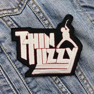 Op de afbeelding: Zwart-witte geborduurde patch met de tekst "Thin Lizzy" in wit met een rode omtrek. Een wit silhouet van een persoon die een gitaar vasthoudt, bevindt zich boven de tekst.