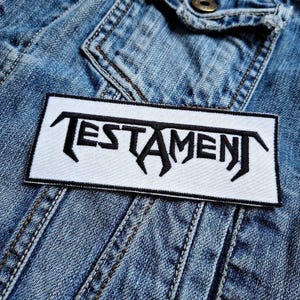 May include: A rectangular white patch with the word "TESTAMENT" embroidered in black, featuring a stylized, gothic font. The patch is placed on a blue denim jacket, showcasing the potential application of the patch.