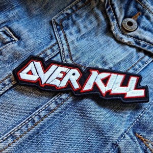 Puede incluir: Parche bordado en blanco y negro con la palabra "OVERKILL" en letras rojas y blancas. El parche está cosido en una chaqueta vaquera azul.