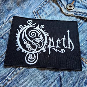 May include: Black embroidered patch with a white design of a stylized circle with swirls and the word "Opeth" in a script font.