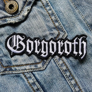 May include: A black and white embroidered patch with the word "Gorgoroth" in a gothic font, sewn onto a blue denim jacket.