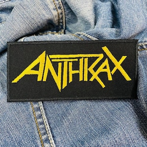 Peut inclure: Un écusson rectangulaire noir avec le mot "ANTHRAX" brodé en jaune. L'écusson est posé sur une veste en jean. Le texte est dans une police audacieuse et stylisée, avec des lettres légèrement inclinées.