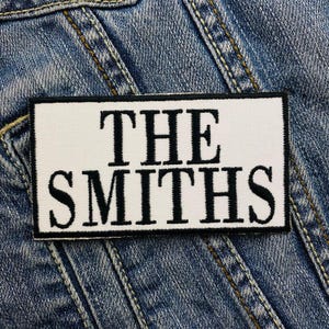 Puede incluir: Parche blanco con texto negro que dice "The Smiths" sobre un fondo de mezclilla azul.