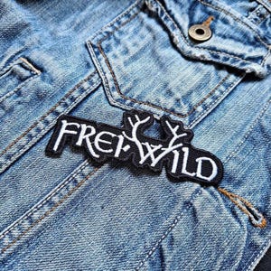 Op de afbeelding: Zwarte geborduurde patch met witte tekst "FREI WILD" en een gestileerde afbeelding van twee geweien.