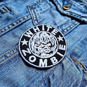 Op de afbeelding: Een geborduurde patch in zwart en wit met de woorden "White Zombie" in een cirkel rond een witte schedel met een dreigende uitdrukking. De patch is genaaid op een blauw denim jack.