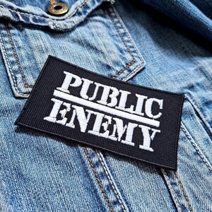 Puede incluir: Parche bordado en blanco y negro con el texto "PUBLIC ENEMY" en una chaqueta vaquera.