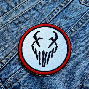 May include: A white and red embroidered patch with a black skull design. The skull has two X's for eyes and a jagged line for a mouth.