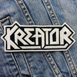 Op de afbeelding: Witte geborduurde patch met het woord "Kreator" in zwart op een denim achtergrond.