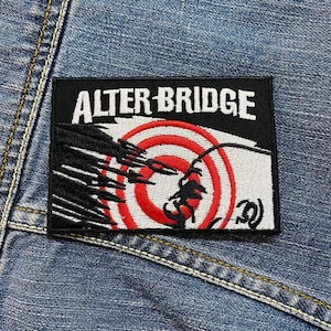 Op de afbeelding: Een rechthoekige zwart-witte patch met de tekst "ALTER BRIDGE" in witte blokletters. De patch heeft een rood-wit targetontwerp met een zwart silhouet. De patch is op een spijkerjas genaaid.