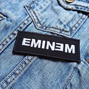 May include: A black patch with white embroidered text that reads "EMINEM" is sewn onto a blue denim jacket.