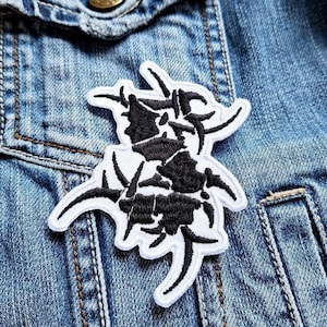 Könnte beinhalten: Schwarz-weiß gestickter Patch mit einem stilisierten Logo mit einem gezackten, abstrakten Design. Der Patch ist auf eine blaue Jeansjacke genäht.