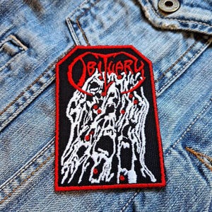 May include: A rectangular patch with a black background and a red border. The word "OBITUARY" is in red, stylized lettering at the top. Below the text is a white and red graphic design. The patch is on a denim jacket.