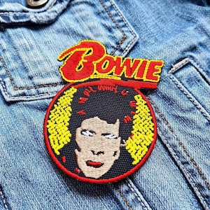 Könnte beinhalten: Ein gestickter Bügel-Patch mit einem Porträt von David Bowie und dem Wort "BOWIE" in roten Buchstaben über seinem Kopf. Der Patch befindet sich auf einem Jeans-Hintergrund.