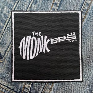 May include: Black square patch with a white border. The patch features the text "THE MONKEES" in a stylized font shaped like a guitar. The patch is likely an accessory for clothing or bags.