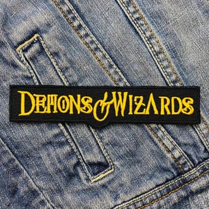 Pode incluir: Etiqueta bordada preta e amarela com o texto "Demons & Wizards" em um fundo de jeans.