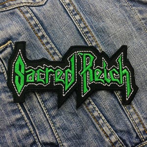 May include: Green and black embroidered patch with the text "Sacred Reich" on a denim background.