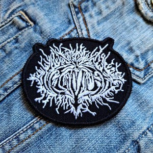 May include: Black and white embroidered patch with a detailed design of a skull with horns and a floral pattern. The patch is sewn onto a blue denim fabric.