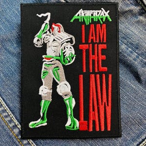 May include: Black and red embroidered patch with a white figure holding a weapon. The text "Anthrax I Am The Law" is embroidered in red.
