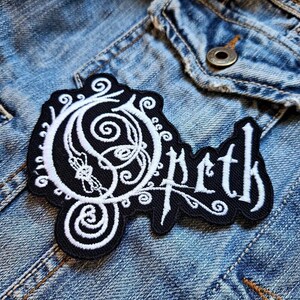 Pode incluir: Etiqueta bordada preta e branca com o nome da banda "Opeth" em uma fonte estilizada. O "O" é um design grande e intrincado com redemoinhos e floreios.