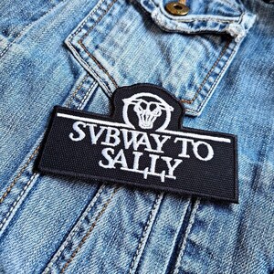 以下が含まれることがあります： 「SUBWAY TO SALLY」の文字とスカルデザインが施された、白と黒の刺繍パッチ。長方形のパッチは白い縁取りがあり、薄い青色のデニムジャケットに取り付けられています。