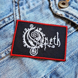 May include: A black and white embroidered patch with a red border. The patch features a stylized logo with the word "Opeth" and a swirling design.