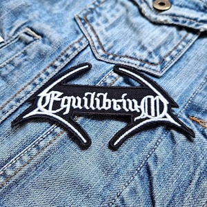 Könnte beinhalten: Ein schwarz-weißer, gestickter Aufnäher mit dem Wort "Equilibrium" in einer stilisierten Schriftart. Der Aufnäher ist auf eine hellblaue Jeansjacke genäht, mit sichtbaren Nähten und einem Knopf.