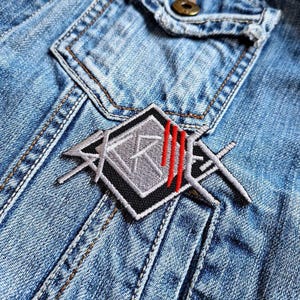 May include: A black and gray embroidered patch with a stylized logo and red stripes. The patch is sewn onto a blue denim jacket.