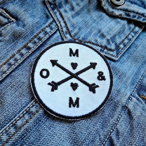 May include: A round, white patch with a black border, featuring crossed arrows, the letters "M" and "O", an ampersand, and heart symbols. The patch is sewn onto a blue denim jacket.