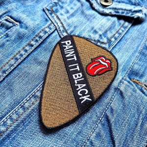Puede incluir: Un parche de púa de guitarra marrón con ribete negro y texto blanco que dice "Paint It Black". Un logotipo de lengua roja está centrado en el parche.