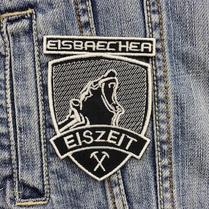 May include: Black and white embroidered patch featuring a wolf howling at the moon. The patch has the text "Eisbaecker Eiszeit" and a pickaxe symbol.