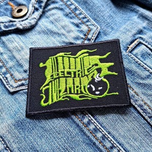 Peut inclure: Un écusson noir brodé avec les mots "ELECTRIC WIZARD" en vert néon, ainsi qu'un dessin de bombe. L'écusson est rectangulaire et est posé sur une veste en jean.