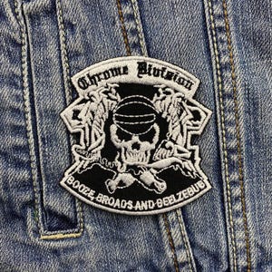 Könnte beinhalten: Ein schwarz-weißer, gestickter Aufnäher mit dem Text "Chrome Division" und "Booze, Broads and Beelzebub". Der Aufnäher zeigt einen Totenkopf, Flügel und andere grafische Elemente, auf eine Jeansjacke genäht.