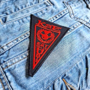 May include: A black triangular patch with red embroidered text that reads "KSE EAT THE DUST". The patch features a stylized skull with a laurel wreath around it.